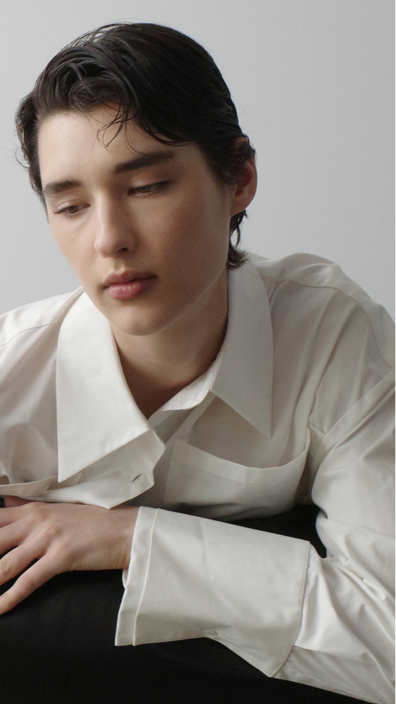 A model wearing a white long-sleeve button-up shirt leans against a black leather sofa against a light gray background.